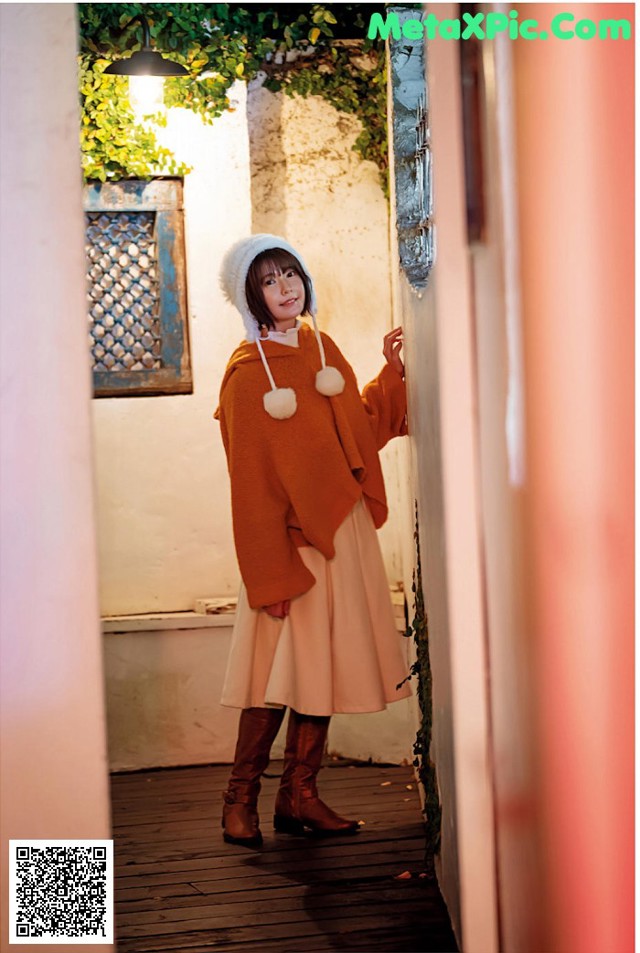 A woman in a brown sweater and a white hat is standing in a doorway.